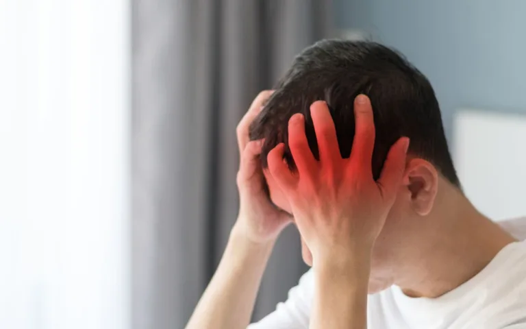 A man with red hair lies in bed, holding his head in pain