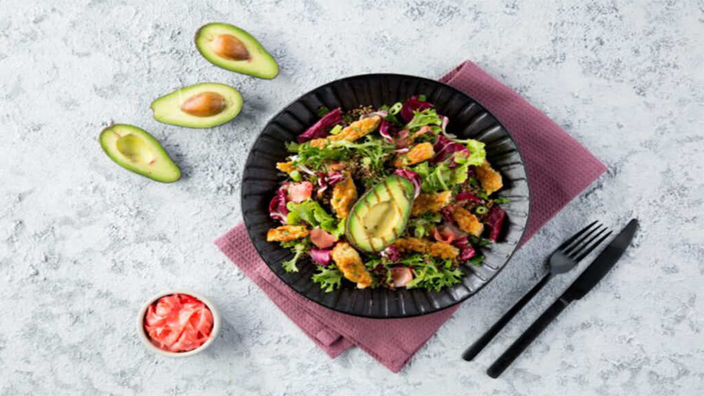Grilled avocado salad with mixed greens, nuts, seeds, and pickled ginger on a black plate.