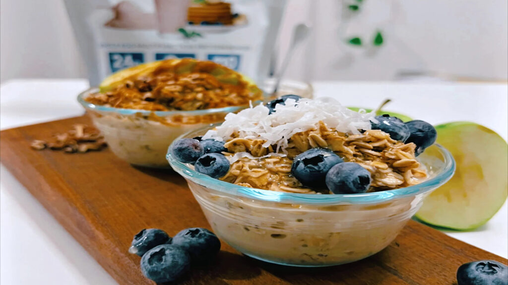 Oatmeal bowls topped with blueberries, coconut, granola, and almond milk in glass bowls on wooden board.