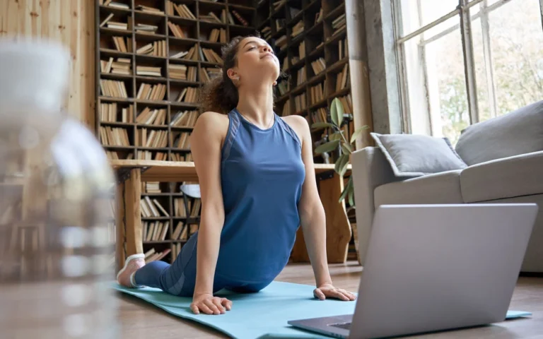 A woman sits on a yoga mat, focused on her laptop