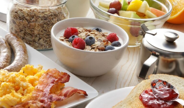 A breakfast spread featuring a bowl of yogurt topped with granola, raspberries, and blueberries, alongside a plate of scrambled eggs, bacon, and sausages, representing potential Low-Sodium Lunch Ideas if portions and ingredients are adjusted. A jar of dry oats, a bowl of mixed fruit, a teapot, and toast with jam are also visible on a light wooden surface.